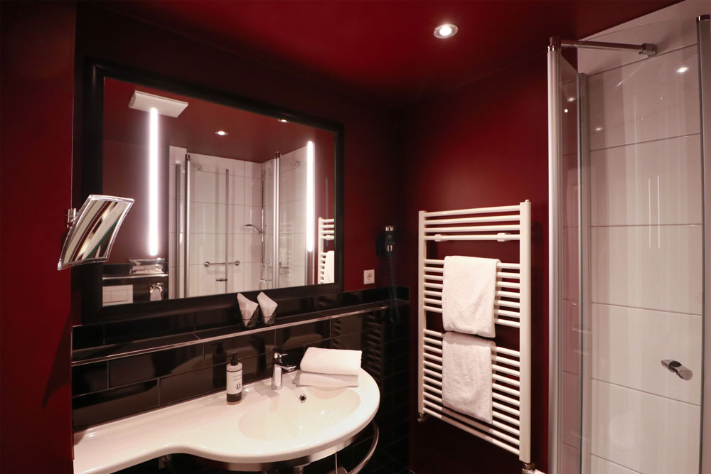 Beleuchtetes Badezimmer mit Waschbecken und Dusche, einem großen Spiegel und einem Heizkörper, den dem zwei Handtücher hängen