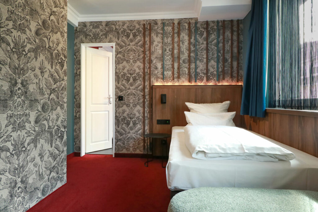 Zimmer mit Einzelbett und gemusterten Tapeten bei Tageslicht