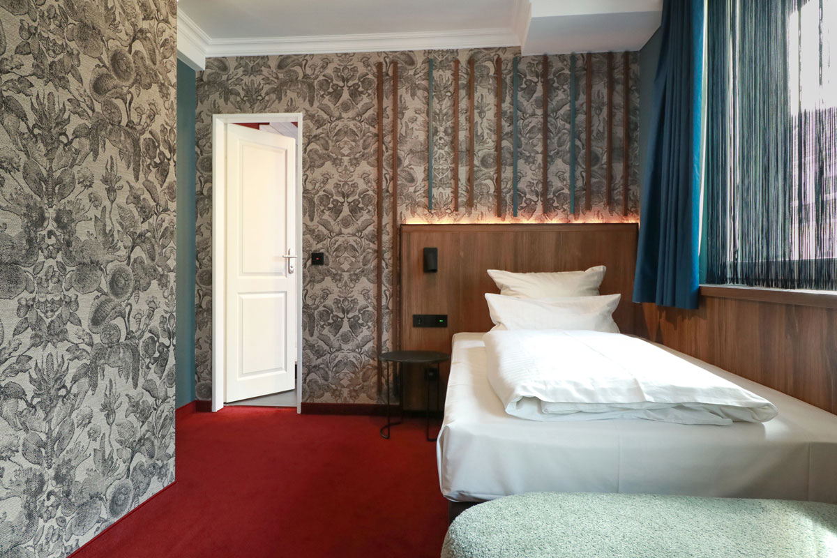 Zimmer mit Einzelbett und gemusterten Tapeten bei Tageslicht