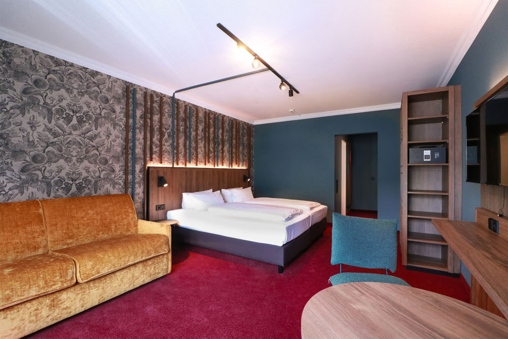 Helles geräumiges Hotelzimmer mit Sofa sowie Doppelbett, Schrank und TV sowie Sitzgelegenheiten