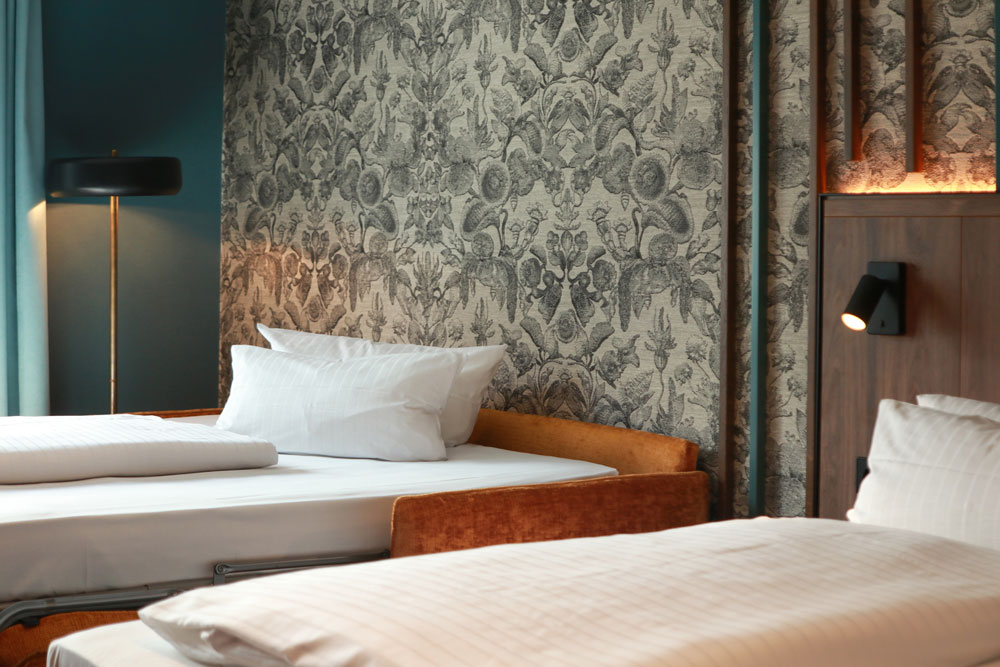 Nahaufnahme eines hellen Zimmers mit aufgeklapptem Schlafsofa und Bett sowie einer Ornament-Tapete