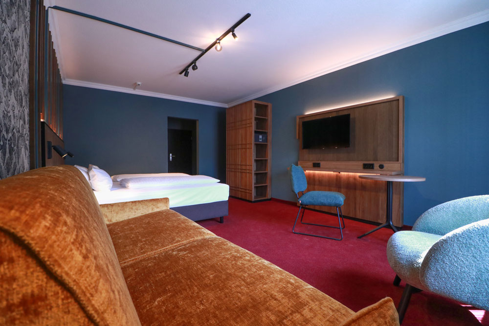 Helles geräumiges Hotelzimmer mit Sofa im Vordergrund sowie Doppelbett, Schrank und TV im Hintergrund