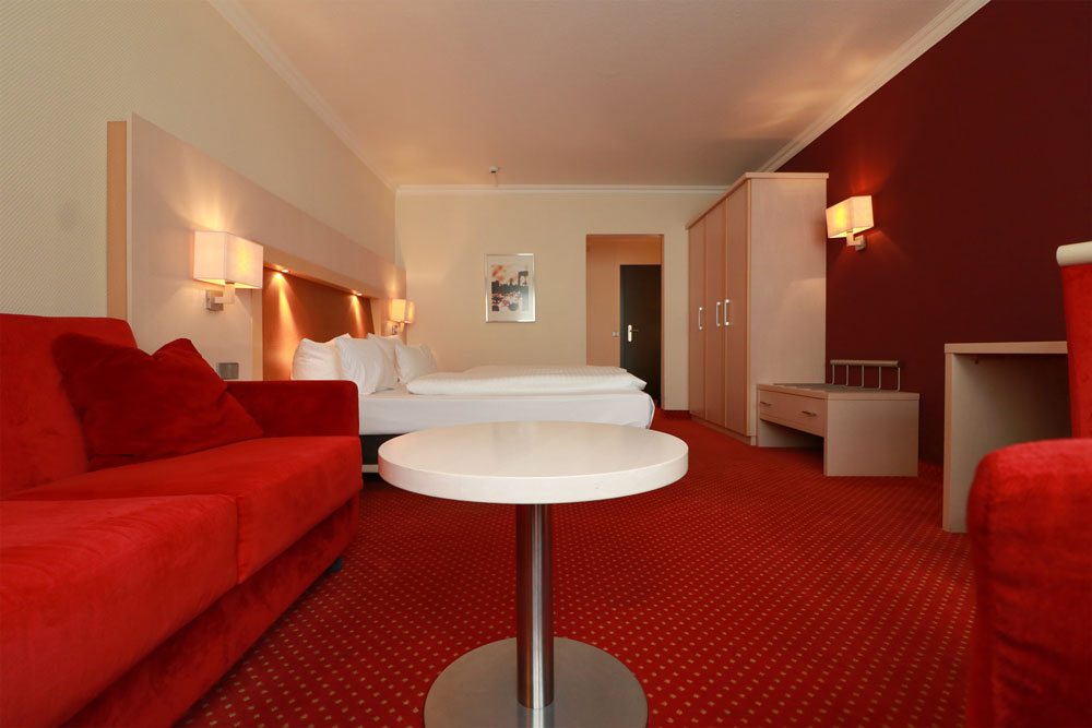Großes Hotelzimmer mit einem Sofa im Vordergrund und einem Doppelbett und Schrank im Hintergrund