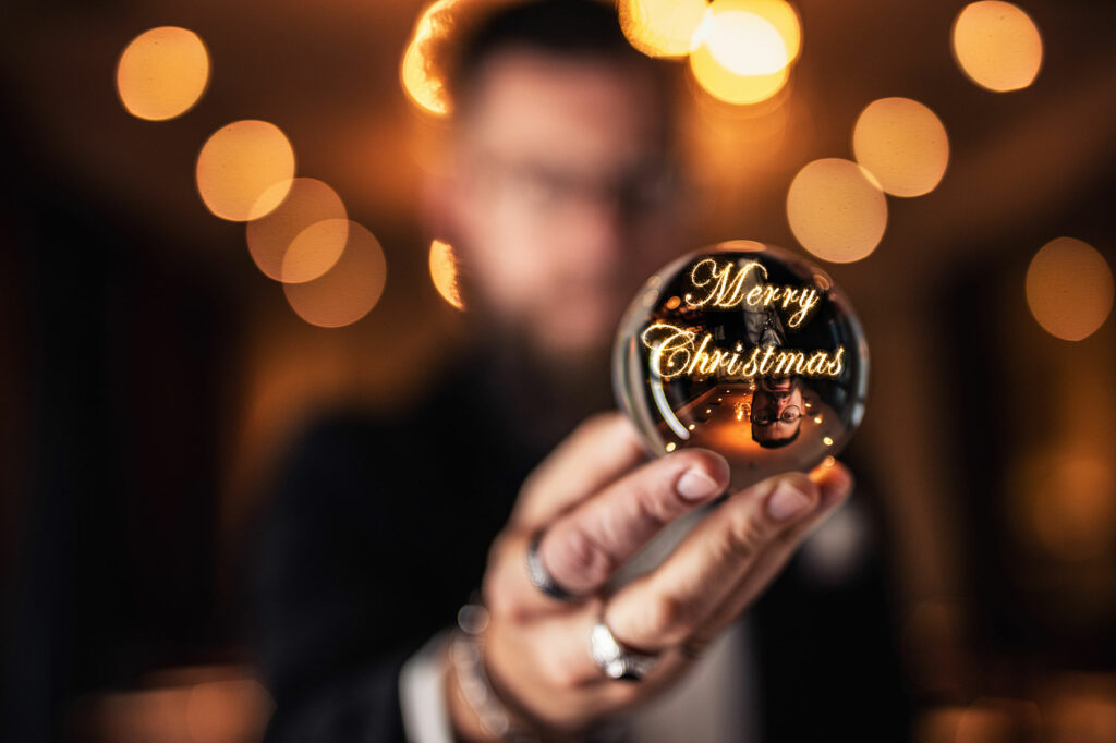 Mann mit einer Glaskugel in der Hand, in der er sich spiegelt und die Wörter Merry Christmas in leuchtender Schrift geschrieben stehen.