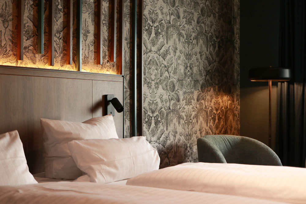 Detailansicht eines Bettes und eines Sessels im Hintergrund, eine Steh- und Bettlampe beleuchten den Raum