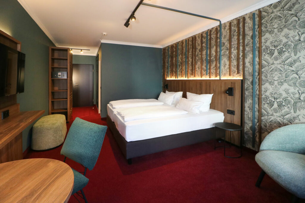 Helles Hotelzimmer mit Doppelbett, Sessel, Tisch, Stuhl, TV-Bank und rotem Teppichboden